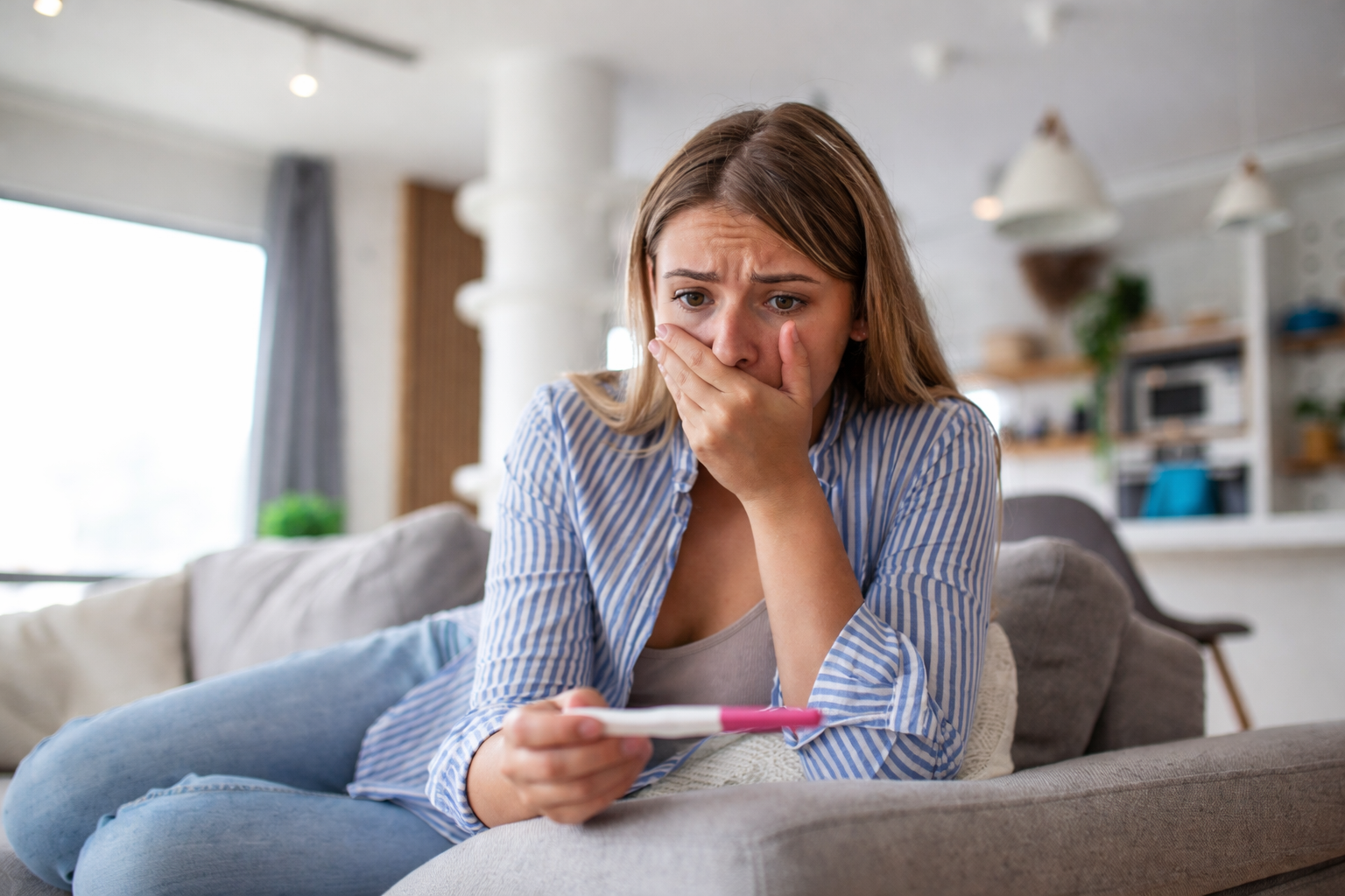 Woman sitting on a couch holding a pregnancy test with a worried expression.