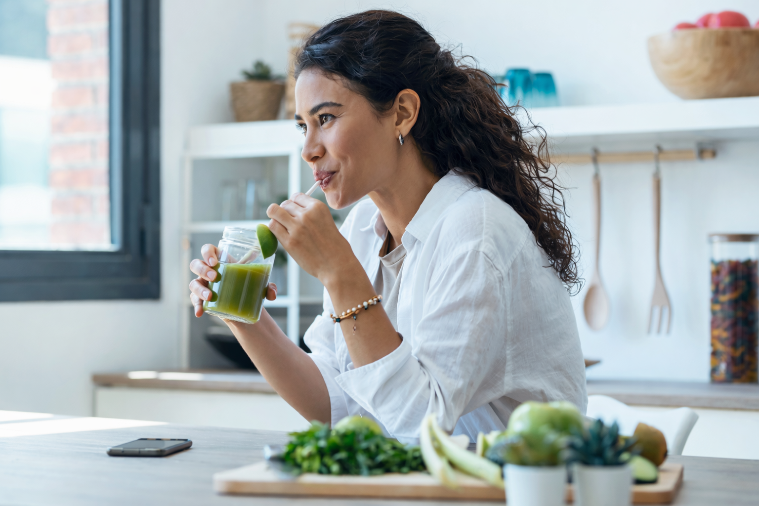 Woman drinking green juice in a kitchen with fruits and vegetables on the counter.