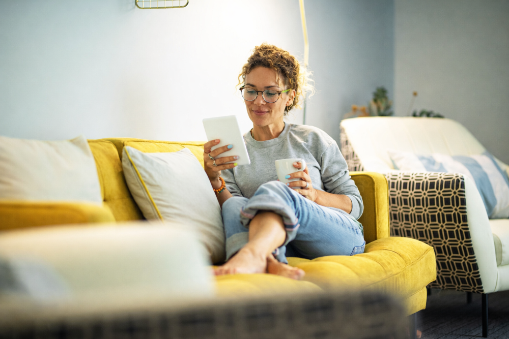 Woman sitting on a yellow couch holding a tablet and a mug in a living room.